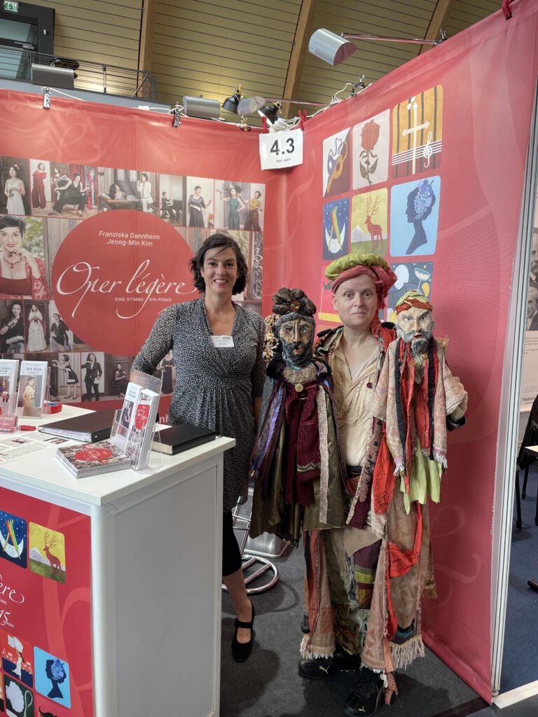 Zwei Frauen an einem Messestand, eine im Theaterkostüm