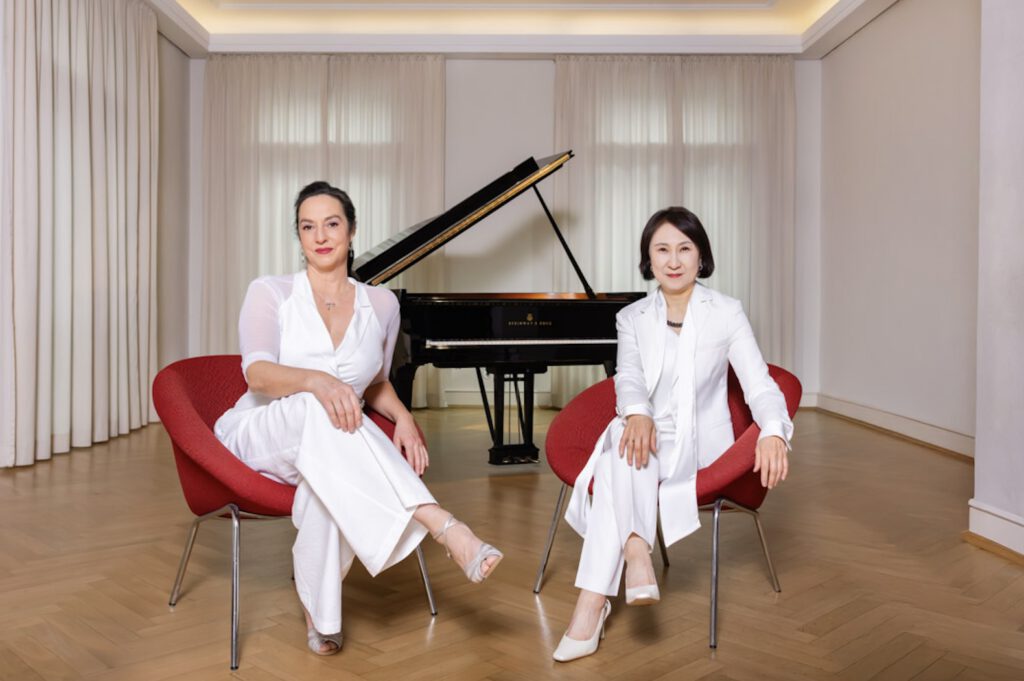 Franziska und Jong-Min sitzen in weißer Kleidung auf roten Sesselchen vor dem schwarzen Flügel