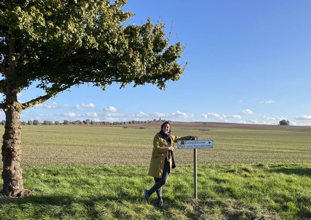 weites Feld, im Vordergrund Franziska Dannheim an ein Wanderschild gelehnt.